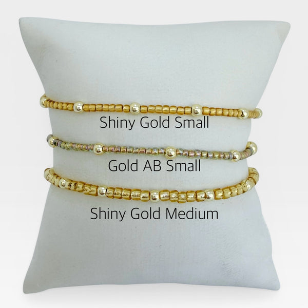 Three gold beaded bracelets on a white cushion with size labels.