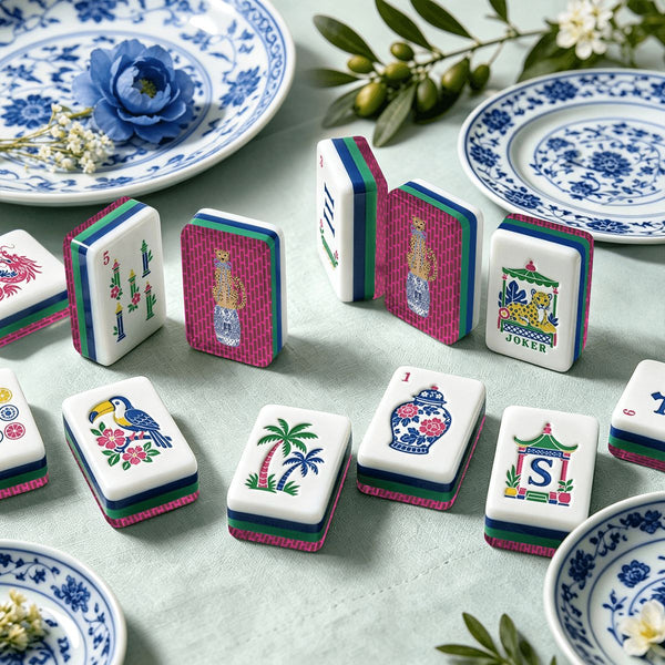 A set of Mahjong tiles arranged on a pale green fabric surface with decorative blue-and-white porcelain plates and greenery in the background. The tiles feature multicolored illustrated faces including birds, palm trees, floral motifs, and traditional symbols.