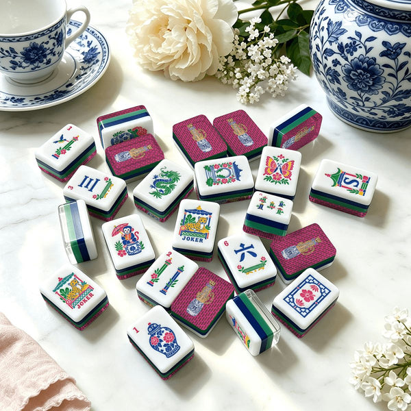 A set of Mahjong tiles arranged on a white marble surface, surrounded by decorative blue-and-white ceramic dishes, flowers, and a pink cloth. The tiles feature multicolored traditional Chinese motifs including dragons, phoenixes, floral patterns, and text such as 'Joker' and 'Three Winds'.