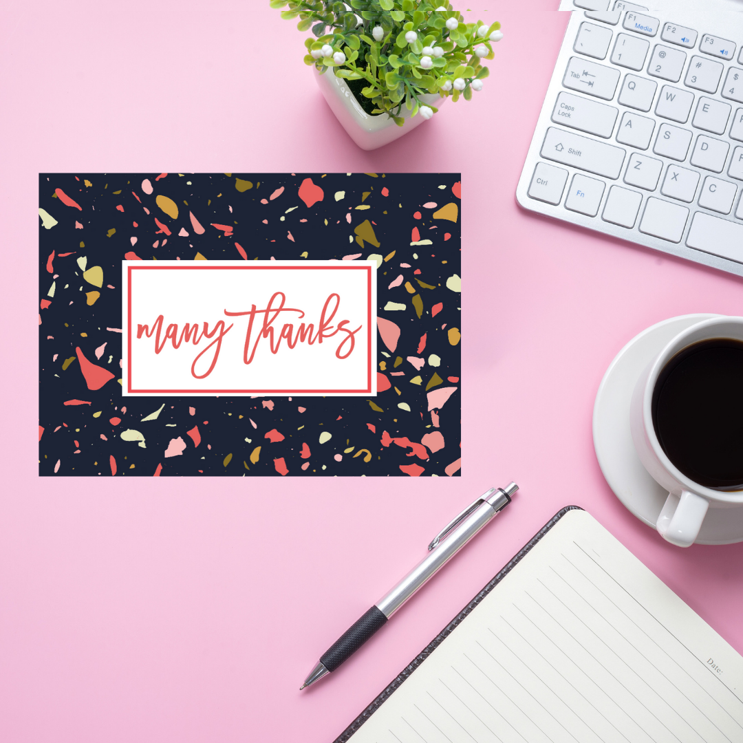 Note card with 'many thanks' on a pink desk with a keyboard, pen, and coffee.