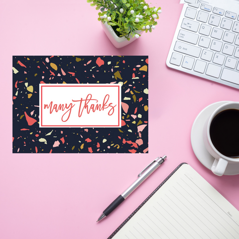 Note card with 'many thanks' on a pink desk with a keyboard, pen, and coffee.