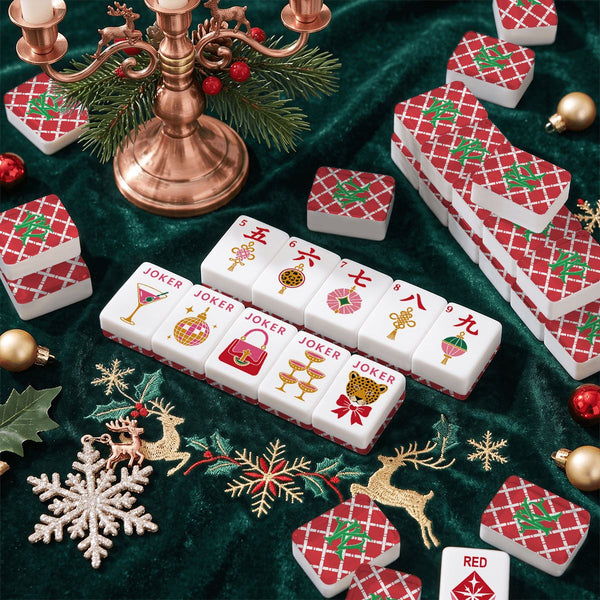 A set of monogrammed Mahjong tiles arranged on a dark green velvet surface with holiday decorations, including a copper candelabra, pine branches, gold ornaments, and embroidered reindeer and snowflake ornaments. The tiles are white with red diamond patterns and colorful printed symbols.