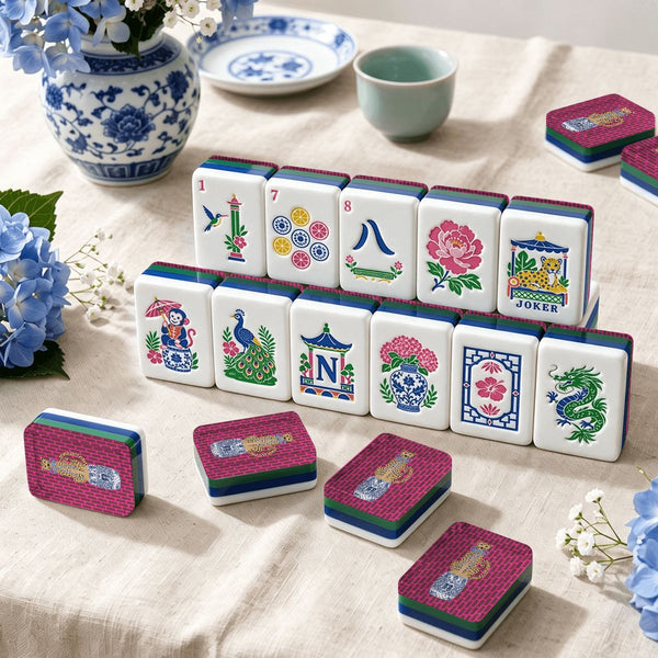 A set of decorative mahjong tiles arranged on a beige fabric surface, with several pink-backed stacks placed around them. The tiles feature colorful Chinese-style floral, bird, dragon, and geometric designs on white faces, with blue and green borders.