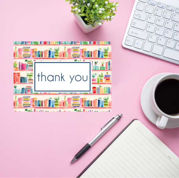 Colorful 'thank you' card with a watercolor bookshelf border on a pink desk with a pen, notebook, coffee cup, and keyboard.