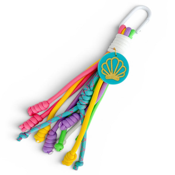 A multicolor paracord keychain tassel with a white carabiner, white cord wrap, a turquoise shell-shaped charm with gold foil detailing, and colorful knotted cords in pink, yellow, purple, green, and blue on a white background.