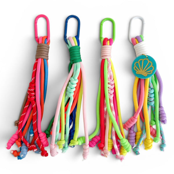 Four colorful carabiner bag charms with textured cords and colorful metal key rings, displayed side-by-side on a white background. One charm has a scallop shell disc.