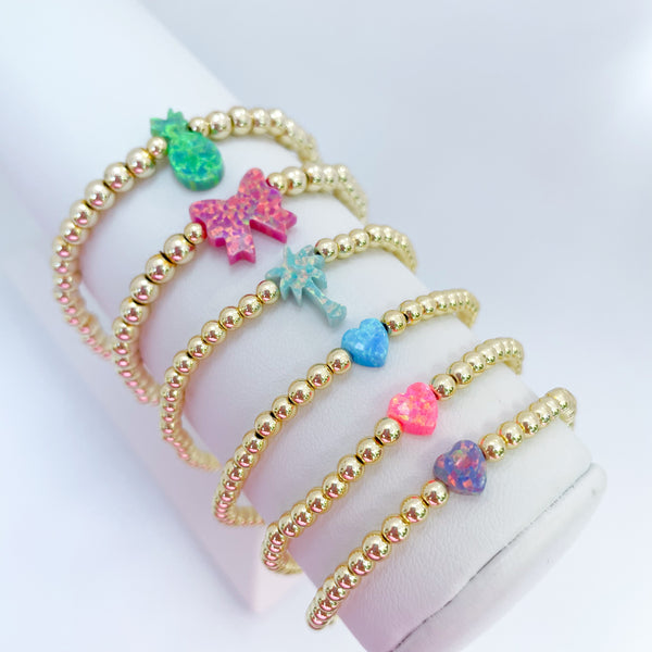 Gold beaded bracelets with colorful opal bow, palm tree, pineapple and heart charms on a white background