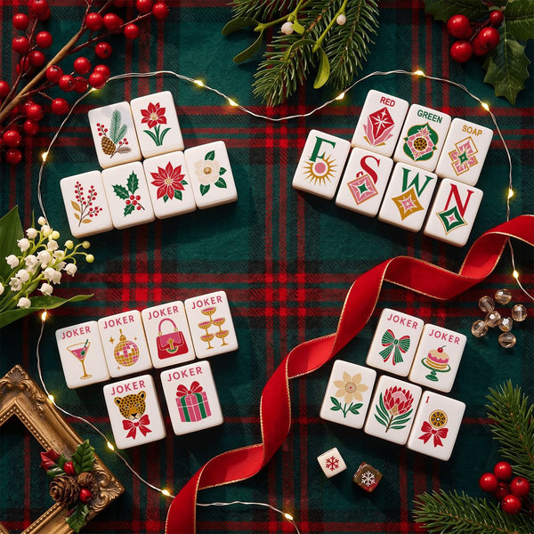 A styled flat-lay of white Christmas-themed jokers and suit tiles arranged on a red and green plaid fabric background with pine branches, berries, fairy lights, and a red ribbon.