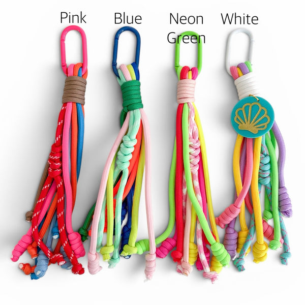 Four colorful braided rope bag charms with hanging loops, labeled Pink, Blue, Neon Green, and White, shown on a plain white background.
