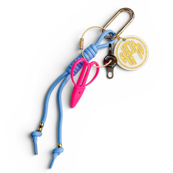 A light blue paracord keychain organizer with gold-tone hardware, featuring pink craft snips, a needle threader, and a round gold monogrammed charm on a white background.