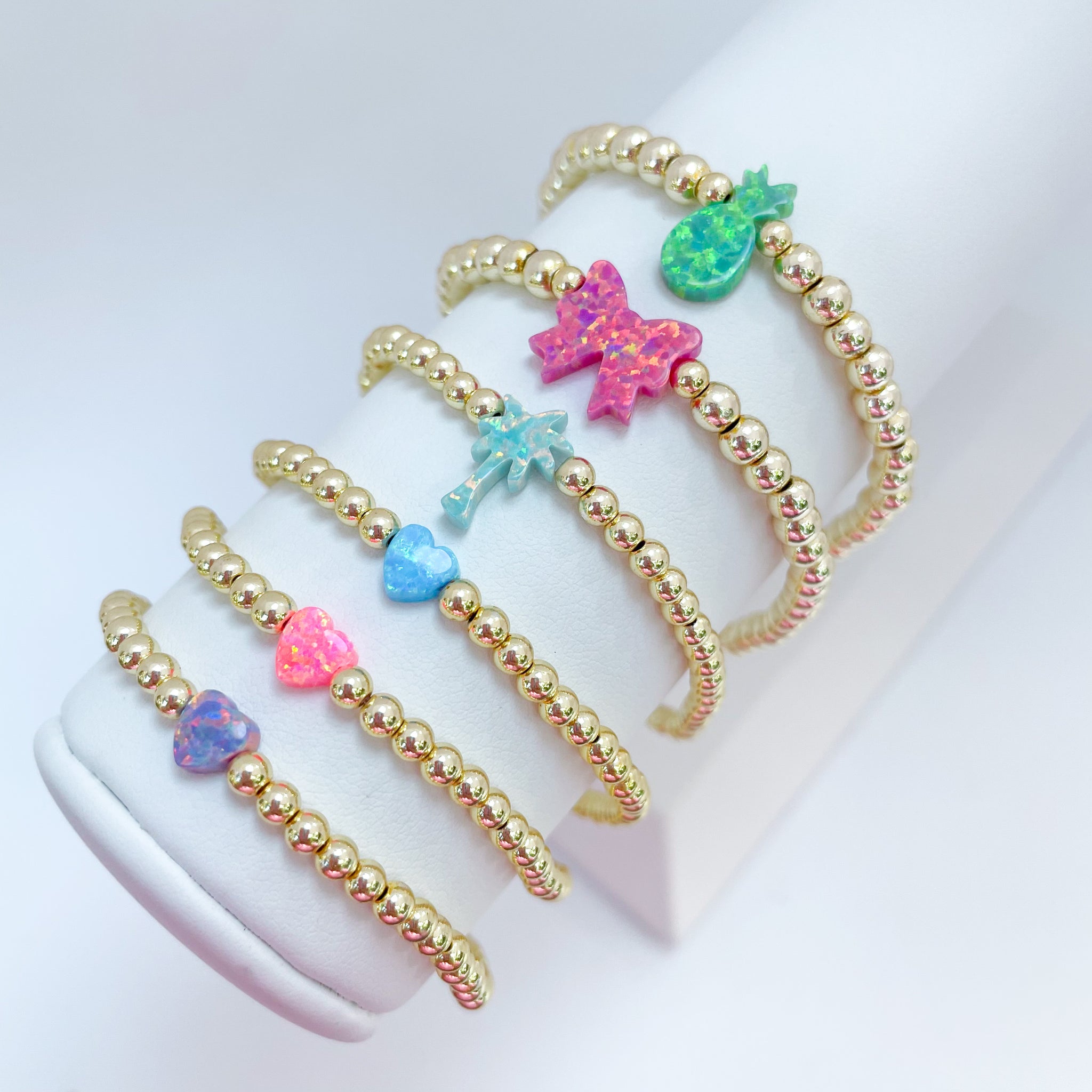Gold beaded bracelets with colorful opal bow, palm tree, pineapple and heart charms on a white background