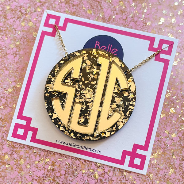 Gold monogram necklace on a branded card with a glittery pink background