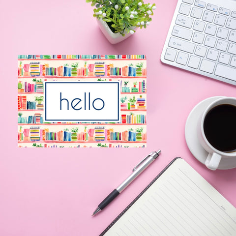 Colorful 'hello' card with a watercolor bookshelf border on a pink desk with a pen, notebook, coffee cup, and keyboard.