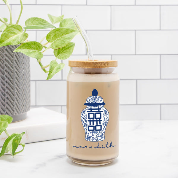 Glass drink tumbler with wooden lid and blue ginger jar on a white surface with plants in the background