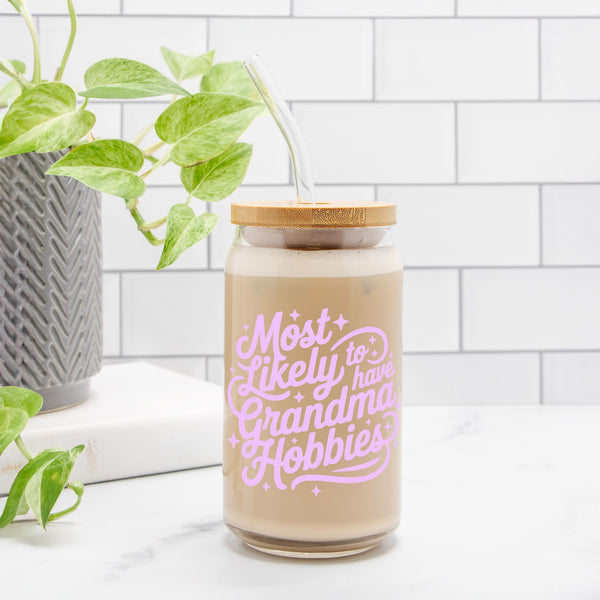 Glass tumbler with straw and wooden lid featuring pink text that says Most Likely to have Grandma Hobbies on a counter with subway tile and a plant