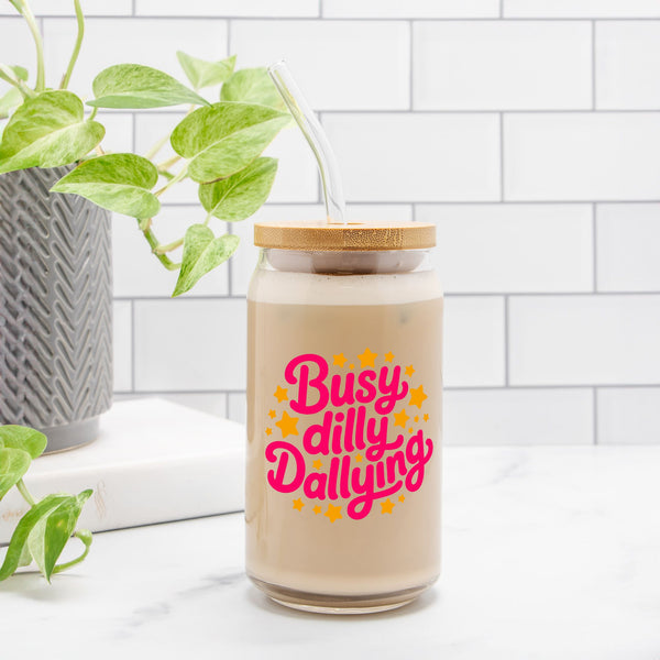 Glass jar with a straw and 'Busy dilly Dallying' text on a white surface with a plant in the background.