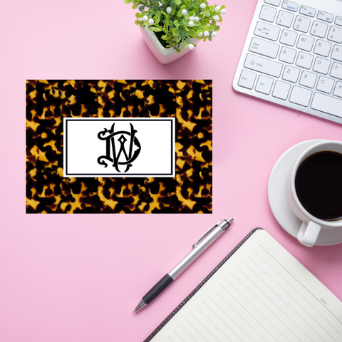 Tortoiseshell print card with monogram on a pink desk with a keyboard, pen, and coffee.