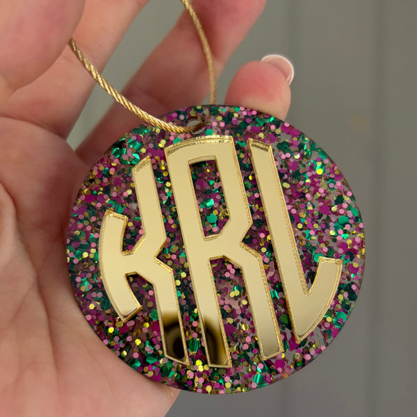 A close-up of a hand holding a round monogram ornament with gold letters 'KRL' over a multicolor glitter background and a gold hanging string.