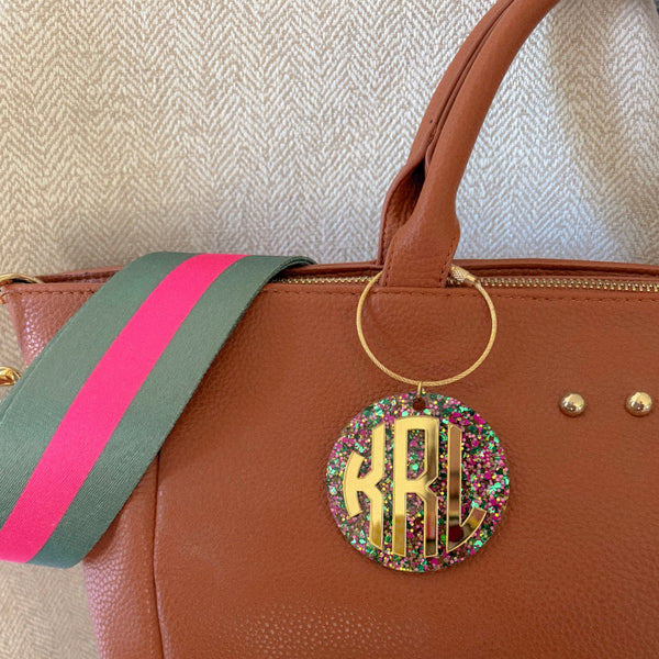 A close-up of a brown leather handbag with a green and pink striped fabric strap, featuring a gold ring and a round glittery monogram charm.