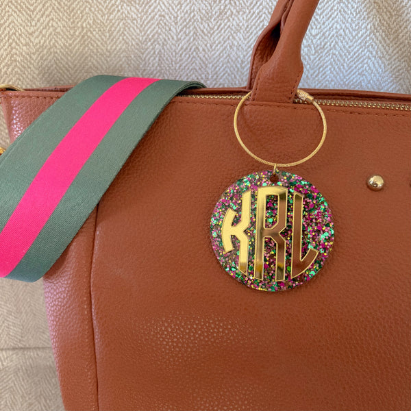 A close-up of a brown textured leather handbag with a colorful striped shoulder strap and a round gold monogram charm covered in multicolor glitter.