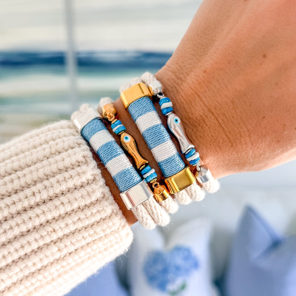 Close-up of a wrist wearing multiple bracelets with blue, white, and gold accents, with a blurred background.