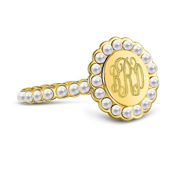 A gold ring featuring a scalloped border of white pearls and a central oval face engraved with an ornate monogram, shown on a clean white background.