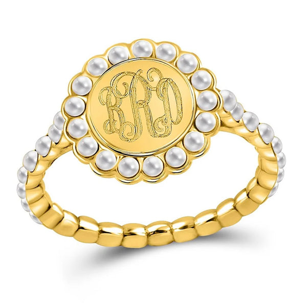 A gold signet ring featuring a round face engraved with an ornate monogram, surrounded by a border of white faux pearls, and a beaded gold band, shown on a white background.