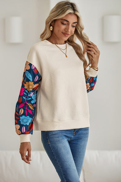 Woman wearing a cream sweater with colorful floral sleeves and blue jeans in a bright room.